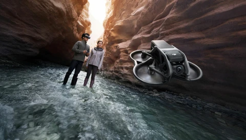 Two people standing in shallow water between towering canyon walls with a drone hovering nearby.