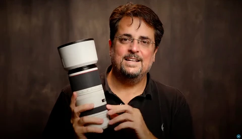 Photographer holding a telephoto lens with white barrel and black grip rings against neutral background.