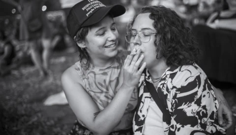 Two people laughing together outdoors in black and white, one wearing a baseball cap and patterned shirt.