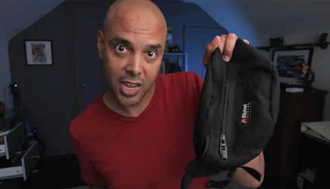 Man in red shirt holding black camera bag toward camera with surprised expression.