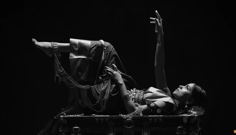 Woman in ornate costume lying on floor with arms extended, shot in dramatic high-contrast black and white.