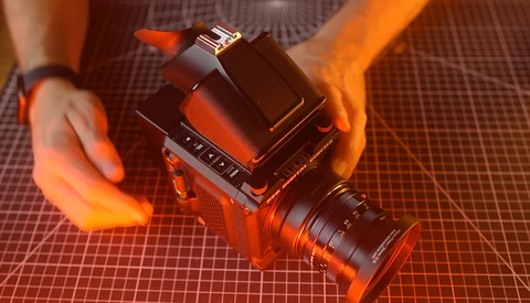 Medium format camera with attached lens held by photographer's hands against orange grid backdrop.