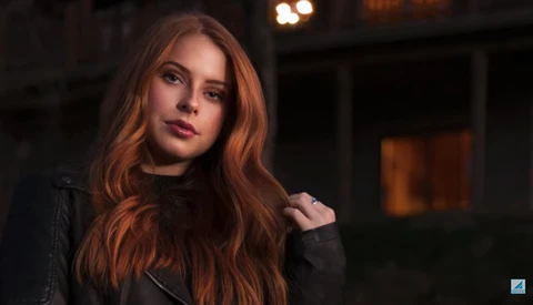 Woman with long reddish-brown hair wearing a black leather jacket, photographed outdoors at night with warm ambient lighting.