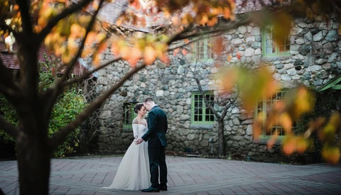 Bride and groom standing together in front of a stone building, framed by autumn foliage.