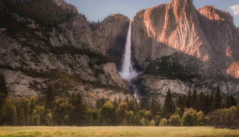 Tall waterfall cascading from granite cliff between steep canyon walls with forested valley floor.