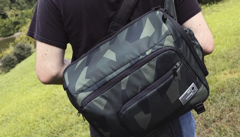 Person wearing a black and gray camo messenger bag with shoulder strap across their chest outdoors.