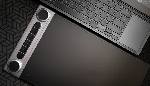 Flat lay of a graphics tablet with stylus pen positioned next to a laptop keyboard on a dark geometric patterned surface.