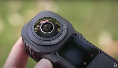 Close-up of a compact action camera with a wide-angle lens held in someone's hand against a blurred green background.