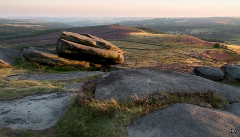Large weathered boulders on a hillside overlooking a patchwork of cultivated fields and distant moorland at golden hour.