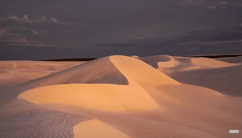 Sand dunes with smooth curves illuminated by golden hour sunlight against a dark stormy sky.