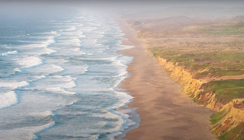 Aerial view of a sandy coastline with white-capped waves and golden cliffs rising above the beach.