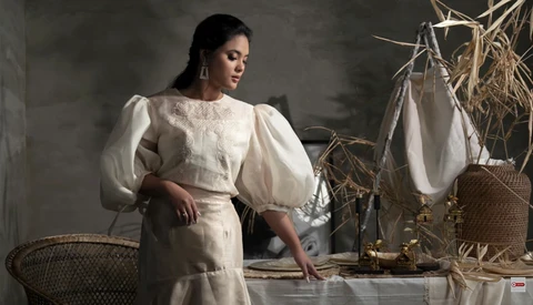 Woman in cream-colored puffed-sleeve dress posed indoors among woven baskets and dried grasses.