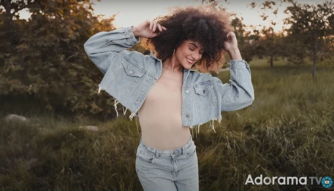 Woman with curly hair posing in an open field at golden hour, wearing an open denim jacket and jeans.