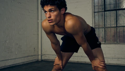 Shirtless man in athletic stance in industrial studio with concrete walls and barred window.