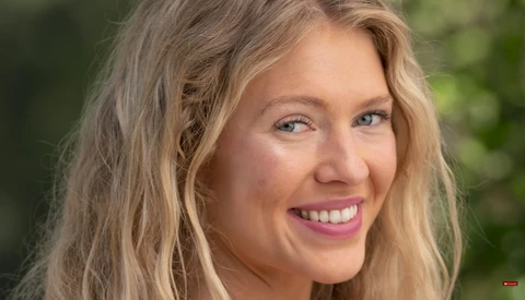 Woman with long blonde wavy hair smiling at camera outdoors with blurred green background.