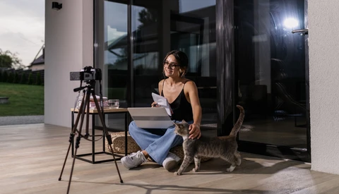 Woman with glasses crouching beside a cat while holding papers, with a camera on tripod and professional lighting setup on a modern patio.