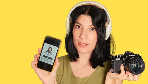 Woman holding smartphone and vintage Nikon camera against yellow background.