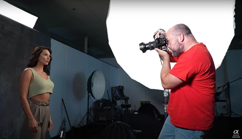 Photographer in red shirt operating cinema camera on tripod while shooting female model in studio with professional lighting setup.