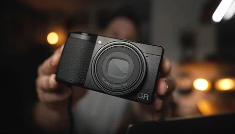 Hands holding a black compact digital camera with prominent lens and GR marking against blurred warm interior lighting.