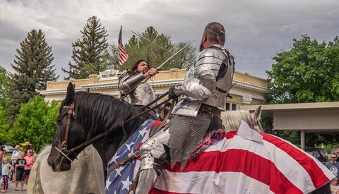 Two people in medieval armor on horseback with American flag draped across the horse.