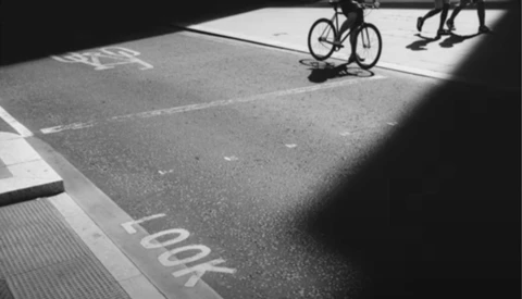High-contrast black and white photograph of a bicycle and pedestrians on a parking lot with dramatic shadows.