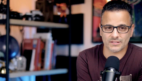 Man wearing glasses and burgundy shirt speaking into a microphone in a home studio setup.