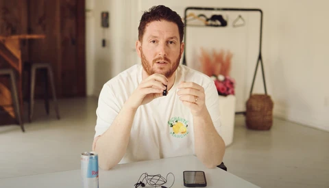 Man in white t-shirt sitting at white table with thoughtful expression, hand near face.