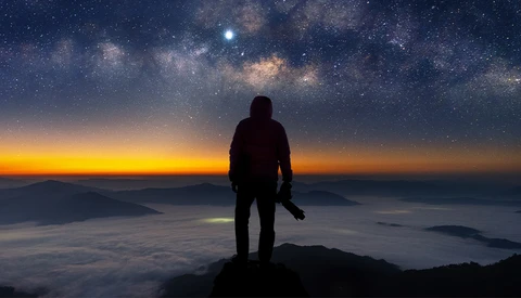 Silhouetted hiker standing on a mountain peak overlooking a sea of clouds at sunrise, with the Milky Way visible in the night sky above.