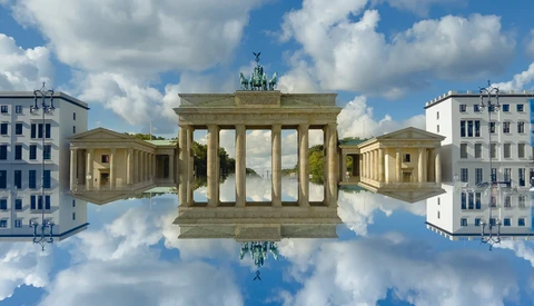 Classical European monument with tall columns reflected in a mirror-like sky effect, flanked by period buildings.