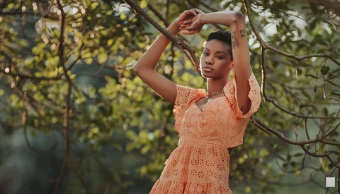 Woman in peach lace dress poses among tree branches with arms raised above head.