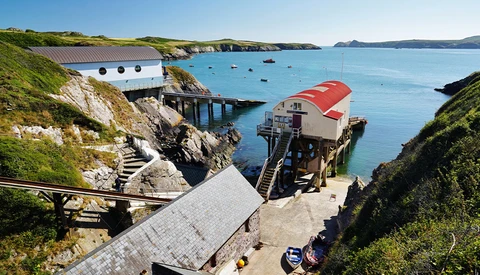Seaside boathouses with red roofs nestled in a cove surrounded by green cliffs and turquoise water.