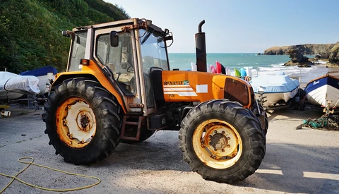 Orange and cream Renault tractor parked at a coastal boat launch, with turquoise water and rocky cliffs visible in the background.