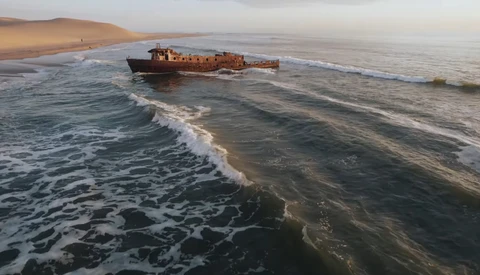 Rusted shipwreck grounded in shallow ocean waters near a sandy desert coastline at sunset.