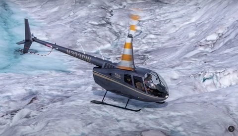 Black and orange helicopter hovering above glacial ice with turquoise meltwater visible at left.