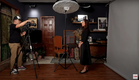 Cameraman on tripod filming a woman in a vintage-styled studio set with warm wood tones and warm lighting.