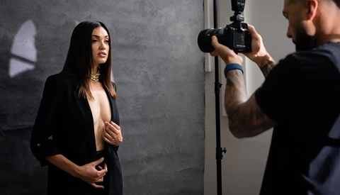 Photographer holding camera on tripod while female subject poses against gray backdrop during studio portrait session.