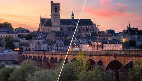 Medieval riverside town at sunset with stone bridge and cathedral spire illuminated by golden and pink sky.