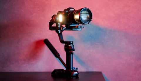 Camera mounted on a three-axis gimbal stabilizer with LED light attached, photographed against a pink and blue gradient background.