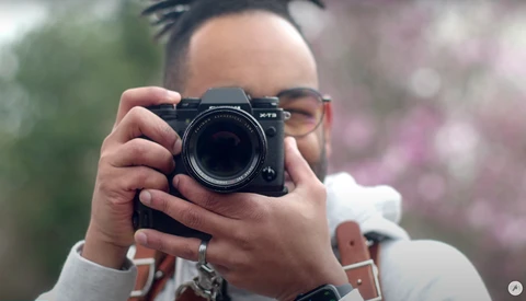 Photographer holding a mirrorless camera up to take a photo, wearing glasses and a white long-sleeve shirt.
