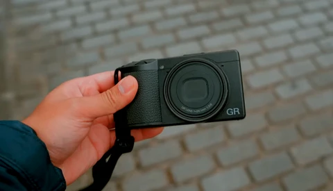 Hand holding a Ricoh GR compact camera against a brick wall background.