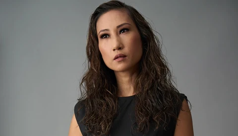 Woman with long dark wavy hair wearing sleeveless black top against neutral gray background.