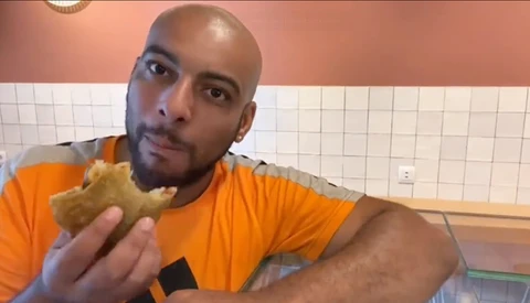 Man in orange shirt holding a sandwich in a tiled kitchen, looking at camera.