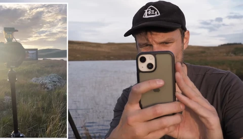 Photographer holding smartphone taking selfie outdoors beside water during golden hour.