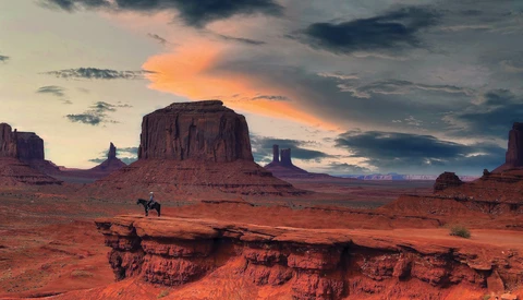 Desert landscape at sunset with rock formations and a lone horseback rider in the foreground.