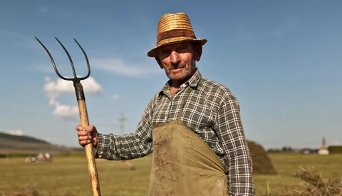 Farmer in straw hat and work apron holding a pitchfork in an open field.