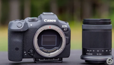 Canon EOS R7 mirrorless camera body with Canon 18-150mm lens displayed against blurred outdoor background.