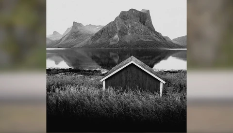 Minimalist black and white landscape with a small wooden boathouse reflected in still water, mountains rising in the background.