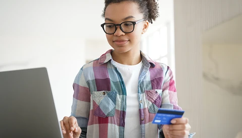 Young person wearing glasses and a plaid shirt holds a credit card while looking at a laptop screen.