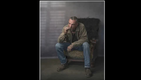 Man in tan jacket sitting on worn upholstered chair against gray wall.