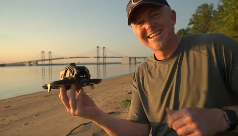 Smiling photographer holding a camera on a beach at golden hour with a suspension bridge visible across the water.